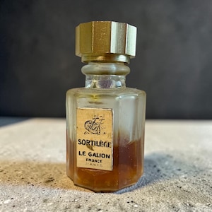 May include: A vintage perfume bottle with a gold-coloured cap and a label that reads "Sortilege Le Galion France Paris." The bottle has a hexagonal shape and contains a dark amber liquid. The label features an illustration of a ship.