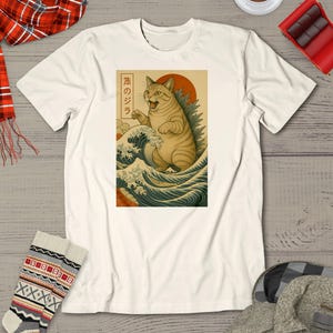 Può includere: T-shirt bianco sporco con una grafica di un gatto in posa Godzilla, ambientata contro un sole che sorge e onde oceaniche. Il design include testo giapponese. Il gatto è marrone chiaro con strisce nere. Le onde sono blu e bianche.