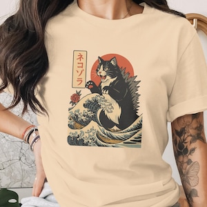 May include: Cream-colored t-shirt featuring a black and white cat illustration in a Godzilla-like pose, set against a red sun and ocean waves. Japanese text is visible on the left side of the graphic. The shirt has short sleeves.