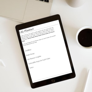 May include: A black tablet displaying a "Life Planner" document with text outlining steps for personal growth, including "Brain Dump" and "Actionable Steps." A cup of coffee and a laptop are visible in the background.