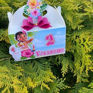 May include: A white party favor box decorated with a pink bow, floral accents, and a cartoon character illustration. The box features the number "2" and the name "Valentina". The box is decorated with a tropical theme.
