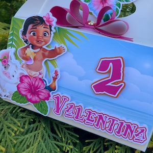 May include: A decorated gift box featuring a cartoon character, a pink bow, and floral accents. The box has the number 2 and the name "Valentina" printed on it. The design includes a tropical theme with a blue sky background.