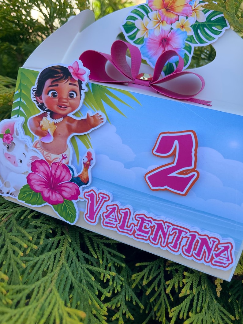 May include: A decorated gift box featuring a cartoon character, a pink bow, and floral accents. The box has the number 2 and the name "Valentina" printed on it. The design includes a tropical theme with a blue sky background.