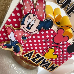 May include: A red and white polka dot cake topper with a Minnie Mouse character, a yellow bow, and the name "Aaliyah" written in black. The topper also includes a red balloon with a yellow Mickey Mouse head.