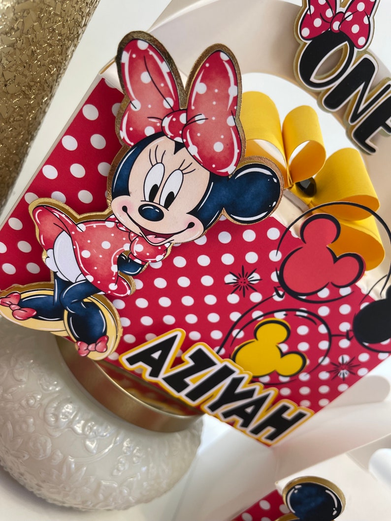 May include: A red and white polka dot cake topper with a Minnie Mouse character, a yellow bow, and the name "Aaliyah" written in black. The topper also includes a red balloon with a yellow Mickey Mouse head.