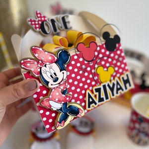 May include: A white favor box with a red and white polka dot pattern. The box features a Minnie Mouse character with a red bow and a yellow bow. The box also has the words "ONE" and "AZIYAH" in gold letters.