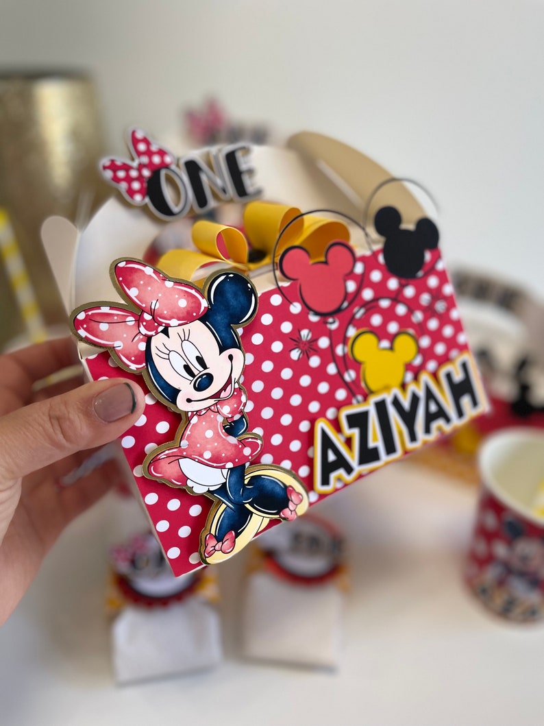 May include: A white favor box with a red and white polka dot pattern. The box features a Minnie Mouse character with a red bow and a yellow bow. The box also has the words "ONE" and "AZIYAH" in gold letters.