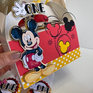 May include: A white and red Mickey Mouse themed party favor box with a gold trim. The box has a handle and says "ONE" on the front. There are two white napkins with red and yellow Mickey Mouse themed napkin rings that also say "ONE" on them.