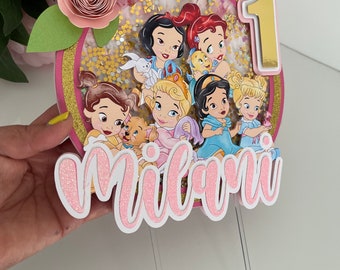 Peut inclure: Un topper de gâteau rose et or avec un cercle représentant sept personnages de princesses Disney, dont Blanche-Neige, Cendrillon et Belle. Le topper comprend également un nom "Milani" en paillettes roses et le chiffre "1" en or.