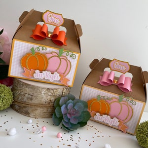 Puede incluir: Dos cajas de regalo marrones decoradas con calabazas naranjas y rosas, un lazo dorado y el texto "One" y "Jai Kyli".