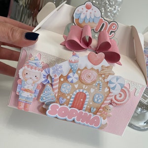 May include: A pink and white favor box with a gingerbread house design. The box has a pink bow and a cupcake topper that says "one". The box also has a nutcracker, a Christmas tree, and the name "Sophia" on it.