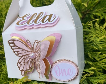 Puede incluir: Caja de regalo blanca con un diseño de mariposa rosa y dorada. La caja tiene un asa y dice "Ella" y "One" en la parte delantera.