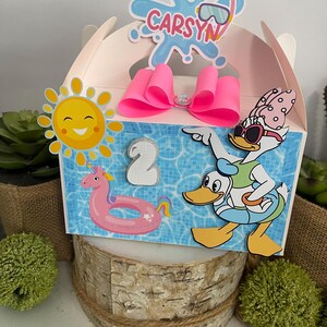 Op de afbeelding: Een lichtroze feestartikelenbox met een zwembadthema. De doos heeft een roze strik, een lachende zon, een eenhoorn drijver en een cartoon eend. De naam "Carsyn" staat bovenop de doos. Het nummer "2" is ook zichtbaar.