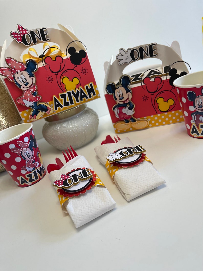 May include: Red and yellow Mickey Mouse and Minnie Mouse themed party favor boxes with the word "One" on the front. The boxes have a handle and are decorated with polka dots. There are also red and yellow cups with Mickey Mouse and Minnie Mouse designs. The cups have the name "Aziyah" on them.