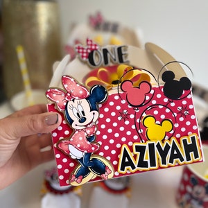 May include: Red and white polka dot favor box with a Minnie Mouse character, a yellow bow, and the name "AZIYAH" in black letters. The box also has a yellow Mickey Mouse head and a red Mickey Mouse head.