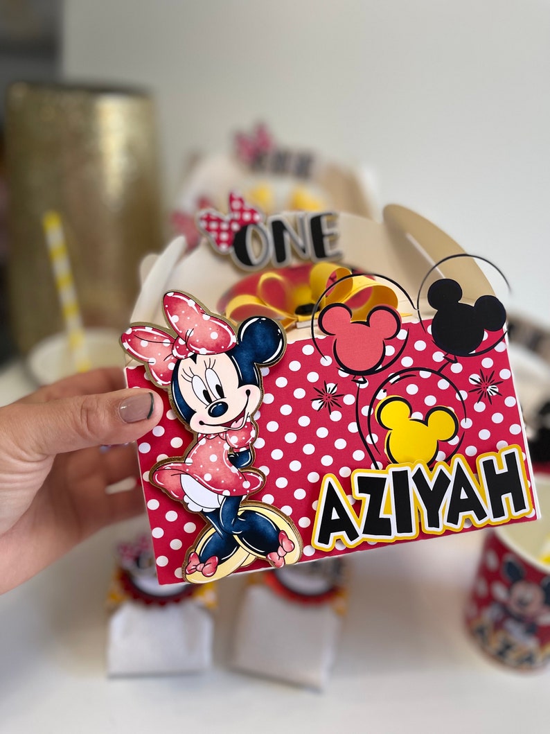 May include: Red and white polka dot favor box with a Minnie Mouse character, a yellow bow, and the name "AZIYAH" in black letters. The box also has a yellow Mickey Mouse head and a red Mickey Mouse head.