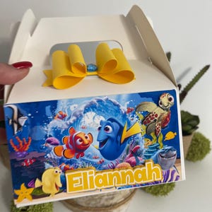 Könnte beinhalten: Eine weiße Geschenkbox mit einer gelben Schleife oben. Die Vorderseite der Box zeigt eine farbenfrohe Unterwasserszene mit Zeichentrickfiguren und dem Namen "Eliannah" in gelben Buchstaben. Die Box ist mit Bildern von Fischen und Meeresbewohnern verziert.