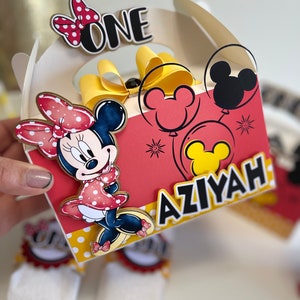 May include: A red and yellow Minnie Mouse themed party favor box with a white handle. The box has a red background with white polka dots and features a Minnie Mouse character, a yellow bow, and the words "ONE" and "AZIYAH".
