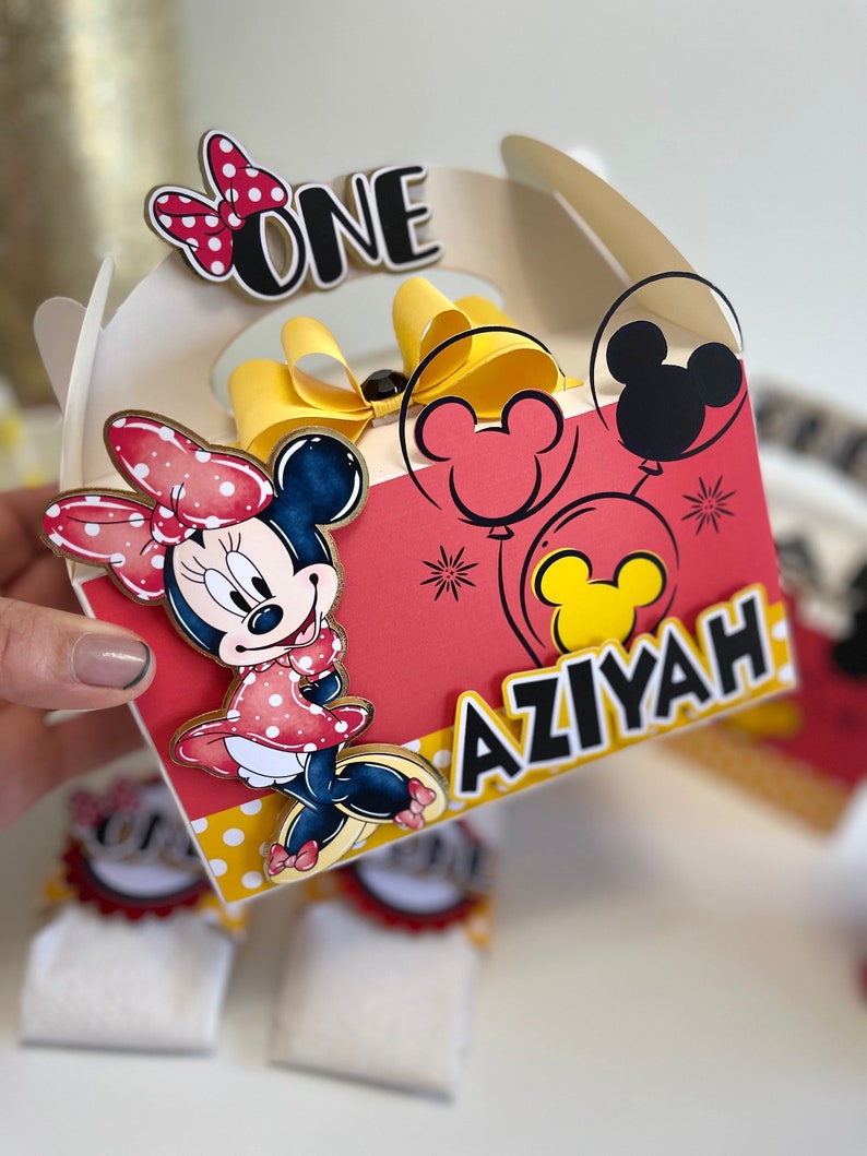 May include: A red and yellow Minnie Mouse themed party favor box with a white handle. The box has a red background with white polka dots and features a Minnie Mouse character, a yellow bow, and the words "ONE" and "AZIYAH".