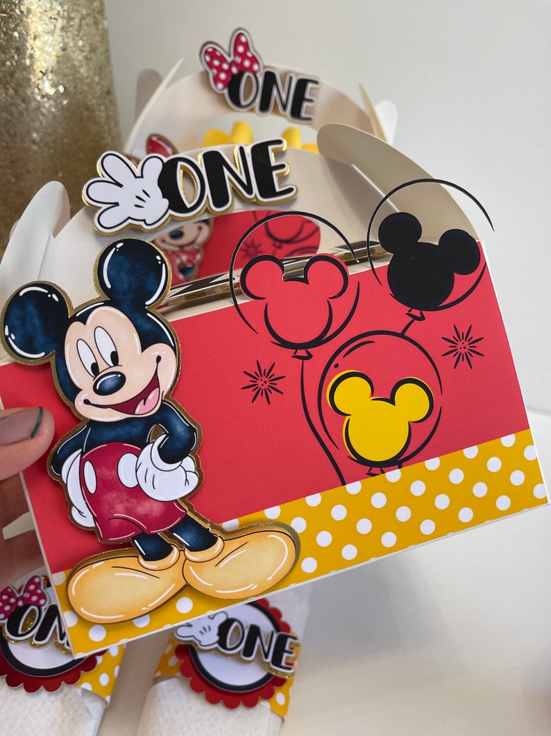 May include: Red and white Mickey Mouse themed party favor boxes with a Mickey Mouse character on the front and the word "ONE" on the top. The boxes have yellow polka dots on the bottom and red and yellow Mickey Mouse balloons on the side.