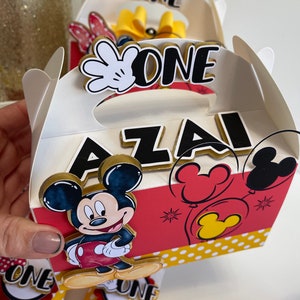 May include: A white favor box with a red and yellow Mickey Mouse design. The box has a handle and says "One" and "Azai" on it.