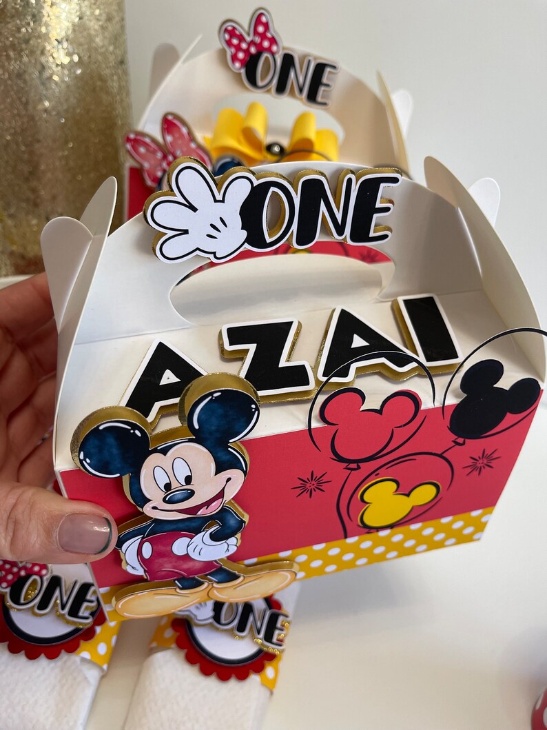 May include: A white favor box with a red and yellow Mickey Mouse design. The box has a handle and says "One" and "Azai" on it.
