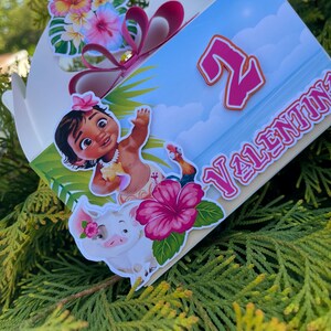 May include: A decorated gift box featuring a cartoon character, a pig, and tropical flowers. The box has a blue background with the number "2" and the name "VALENTINA" in pink. A pink bow and additional floral decorations adorn the top.