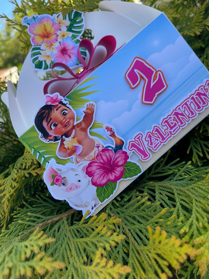 May include: A decorated gift box featuring a cartoon character, a pig, and tropical flowers. The box has a blue background with the number "2" and the name "VALENTINA" in pink. A pink bow and additional floral decorations adorn the top.