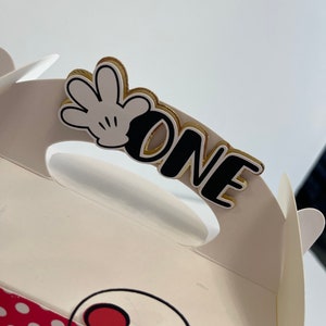 May include: White favor box with a red and white polka dot pattern. The box has a handle with a black and white Mickey Mouse glove and the word "ONE" in black with a gold outline.