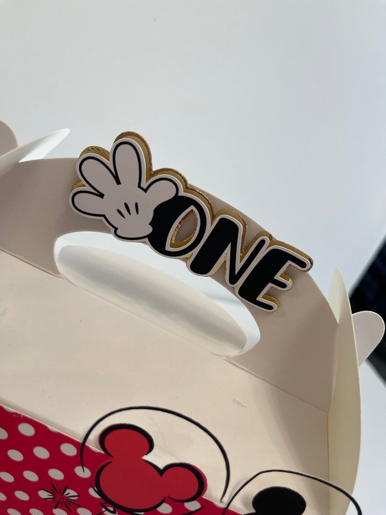 May include: White favor box with a red and white polka dot pattern. The box has a handle with a black and white Mickey Mouse glove and the word "ONE" in black with a gold outline.