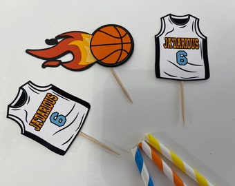 May include: Three cupcake toppers featuring a basketball, a basketball jersey with the name "JADARIUS" and the number "6" in orange and blue, and a second basketball jersey with the same name and number. The toppers are on wooden sticks.