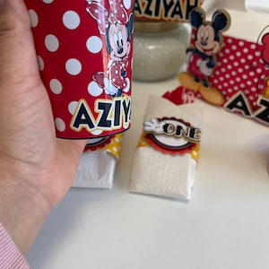 May include: Red and white polka dot paper cup with a Minnie Mouse character and the name "AZIYAH" printed on it. The cup is sitting on a white surface.