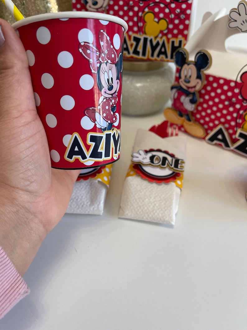 May include: Red and white polka dot paper cup with a Minnie Mouse character and the name "AZIYAH" printed on it. The cup is sitting on a white surface.