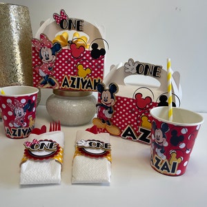 May include: Mickey Mouse and Minnie Mouse themed party supplies, including red and white polka dot cups, favor boxes, and napkin rings with the word "ONE" on them. The boxes have the name "AZIYAH" on them.