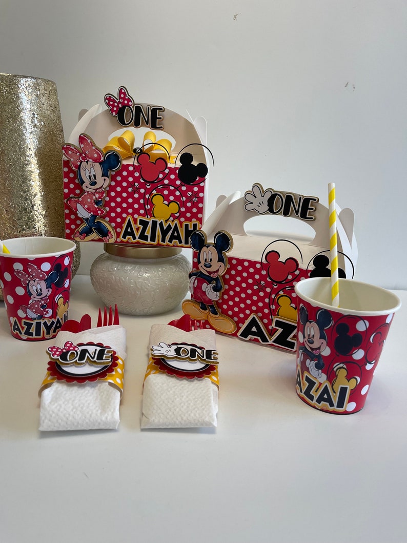 May include: Mickey Mouse and Minnie Mouse themed party supplies, including red and white polka dot cups, favor boxes, and napkin rings with the word "ONE" on them. The boxes have the name "AZIYAH" on them.