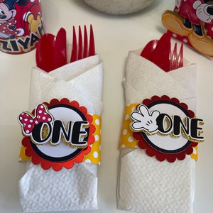 May include: Two white napkins with red and yellow polka dot paper bands wrapped around them. The bands have a red circle with a black outline and the word "ONE" in gold letters. One band has a red bow with white polka dots and the other has a black Mickey Mouse hand. The napkins are folded to hold red plastic forks and spoons.