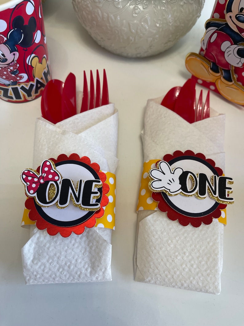 May include: Two white napkins with red and yellow polka dot paper bands wrapped around them. The bands have a red circle with a black outline and the word "ONE" in gold letters. One band has a red bow with white polka dots and the other has a black Mickey Mouse hand. The napkins are folded to hold red plastic forks and spoons.