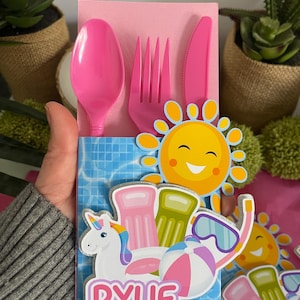 May include: Pink plastic cutlery set with a pink napkin, a sun, a unicorn, and a pool float. The cutlery set is in a pink and blue paper bag with the name "Rylie" written on it.