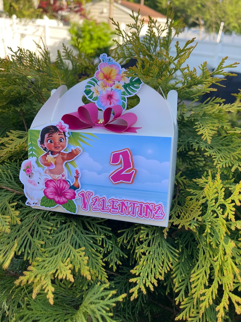 May include: A white gift box decorated with a cartoon image of a girl, a pink flower, and the name "Valentina". The box has a pink bow and a floral arrangement on top. The number "2" is also displayed.