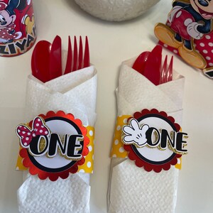 May include: Two red and white Mickey Mouse themed napkin rings with yellow polka dots. One ring has a red bow and the other has a white glove. Both rings have the word "ONE" in gold letters.
