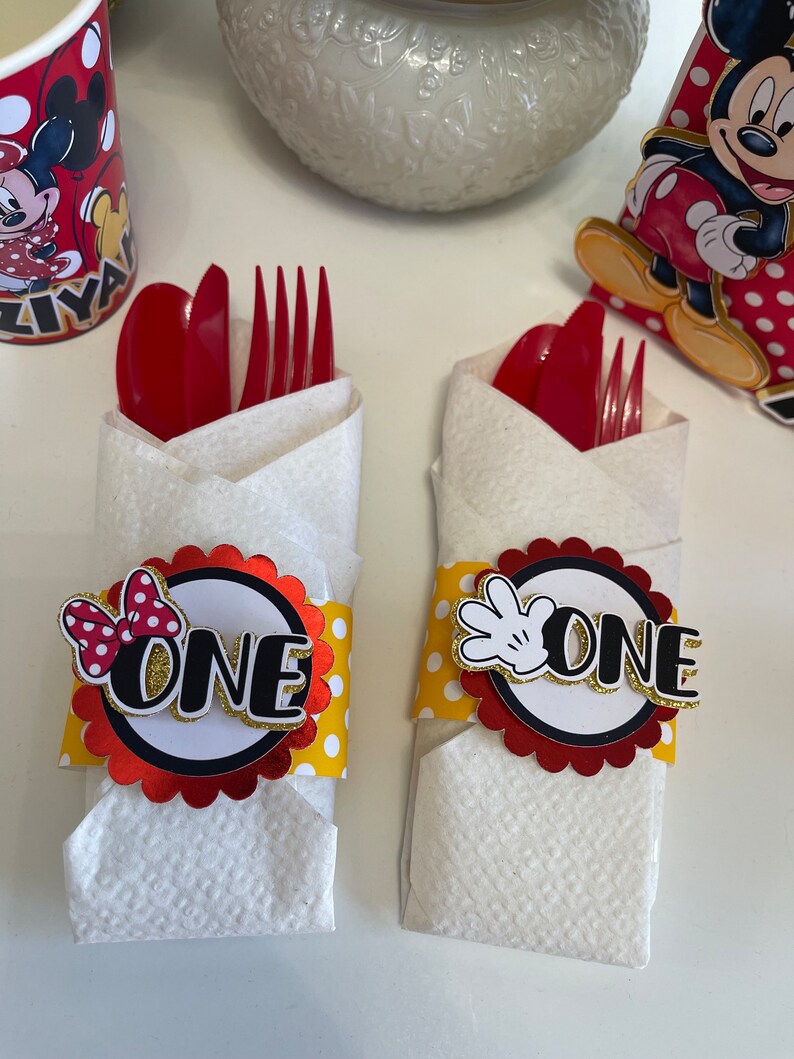 May include: Two red and white Mickey Mouse themed napkin rings with yellow polka dots. One ring has a red bow and the other has a white glove. Both rings have the word "ONE" in gold letters.