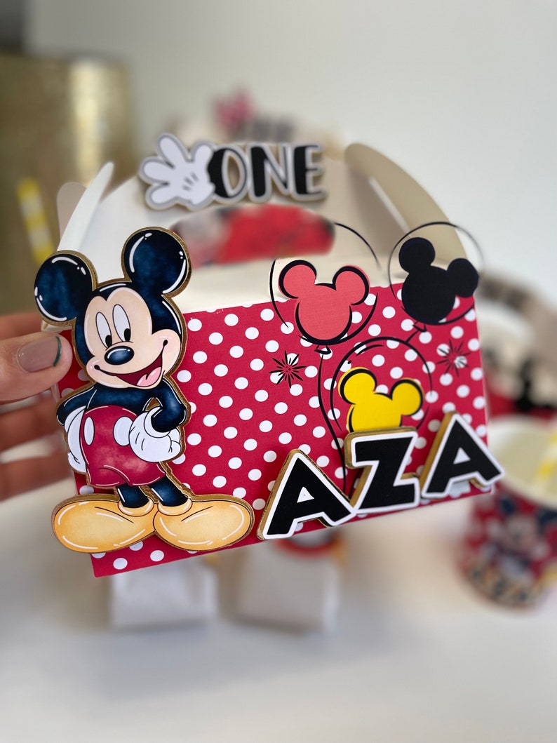 May include: A red and white polka dot favor box with a Mickey Mouse character on the front. The box has a black and white "ONE" on the top and a black "AZA" on the side.