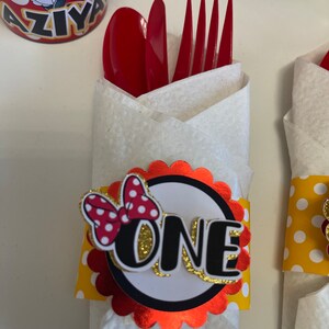 May include: Red and white polka dot bow with a gold glitter "ONE" on a red and white scalloped circle. The circle is attached to a yellow and white polka dot napkin ring. The napkin ring is wrapped around a white napkin with a red fork and spoon.