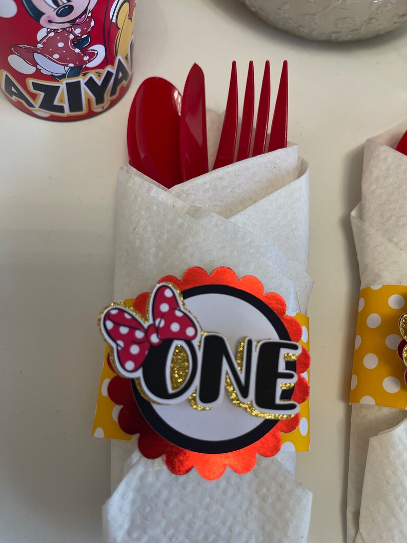 May include: Red and white polka dot bow with a gold glitter "ONE" on a red and white scalloped circle. The circle is attached to a yellow and white polka dot napkin ring. The napkin ring is wrapped around a white napkin with a red fork and spoon.