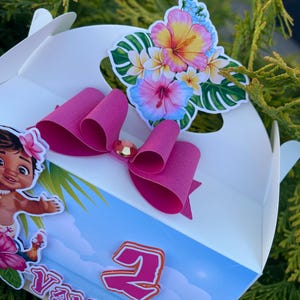 May include: White party favor box with a pink bow and a floral sticker. The sticker features a variety of flowers in pink, yellow, and blue. A cartoon character and the number 2 are also visible on the box.