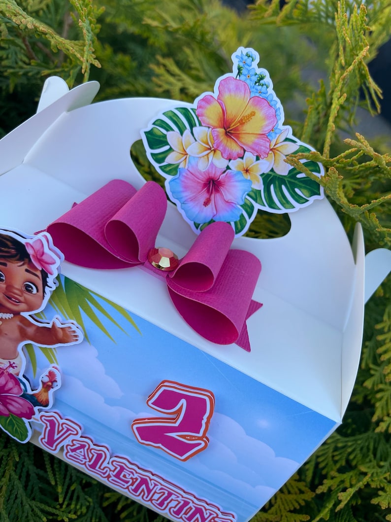 May include: White party favor box with a pink bow and a floral sticker. The sticker features a variety of flowers in pink, yellow, and blue. A cartoon character and the number 2 are also visible on the box.