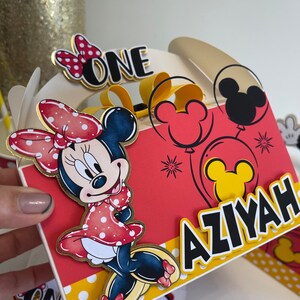 May include: A red and yellow party favor box with a Minnie Mouse character, balloons, and the text "ONE" and "AZIYAH".