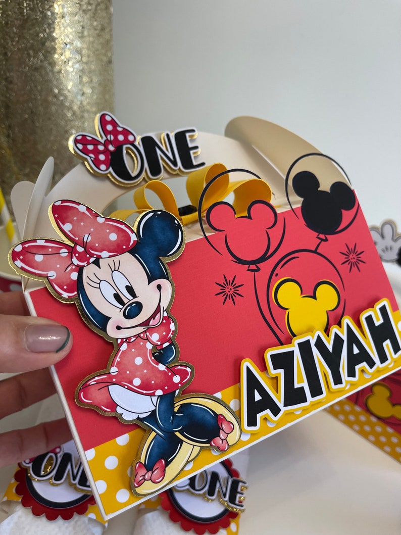 May include: A red and yellow party favor box with a Minnie Mouse character, balloons, and the text "ONE" and "AZIYAH".