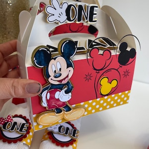May include: A white favor box with a red, yellow, and black Mickey Mouse design. The box has a handle and says "ONE" on the front. The box is decorated with red and yellow polka dots.