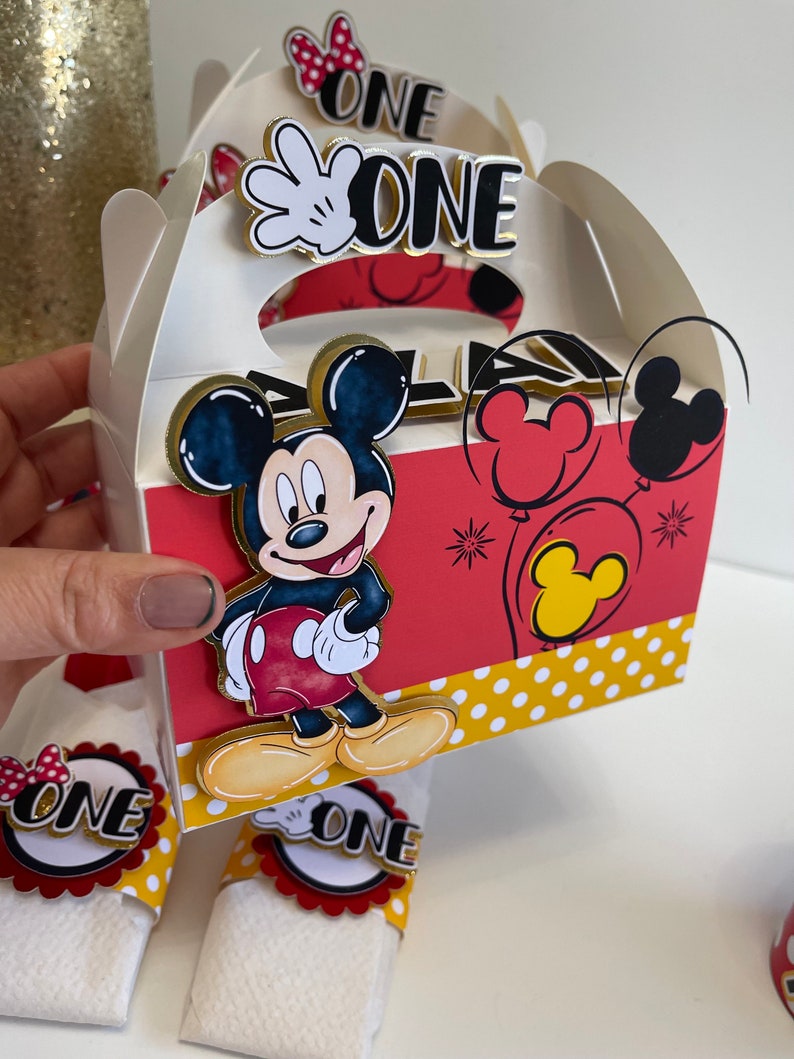 May include: A white favor box with a red, yellow, and black Mickey Mouse design. The box has a handle and says "ONE" on the front. The box is decorated with red and yellow polka dots.